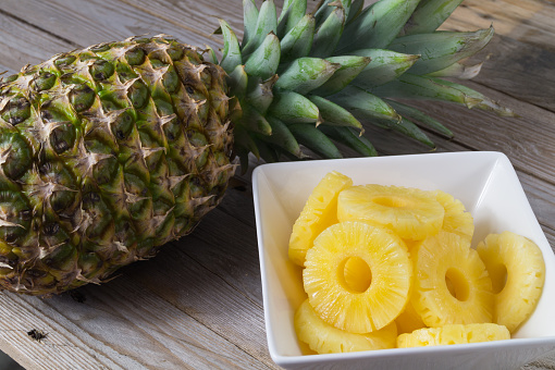 Sliced pineapple kept on a wooden table besides a glass filled with pineapple juice and a glass bowl with pineapple slices