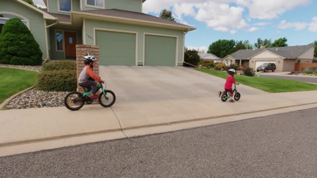Two Caucasian Boys (Five Years-Old and Four Years-Old) Wearing Bike Helmets Ride their Bikes through a Residential Neighborhood under a Partly Cloudy Sky