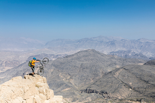 A lonely male mountainbiker is posing at the very edge of a hughe ravine in the desert of the Hajar Mountains which is the highest mountain range in the eastern Arabian peninsula also known as \