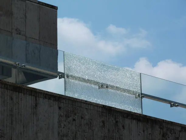 shattered glass railing panel above stone panel under blue sky and white clouds. broken laminated tempered safety glass. construction and building industry concept. 
stainless steel glass brackets.