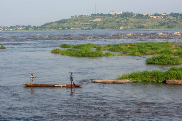 Fishermen paddling between Brazzaville and Kinshasa on Congo River Brazzaville, Republic of Congo - May 20, 2019: Two Congolese men paddling in a typical dugout canoe (pirogue) on Congo River, seen from the shoreline of Brazzaville, Republic of Congo. In the background the Livingstone Falls (also Djoué Falls, a series of rapids between Brazzaville/Kinshasa and Matadi) could be seen. The shoreline in the background is already in the DR of Congo (Kinshasa) and the large building in the background is the Presidential Palace in Kinshasa. kinshasa stock pictures, royalty-free photos & images