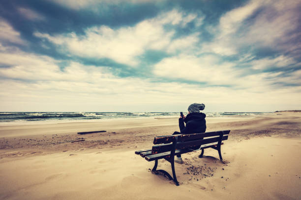 Woman sitting on a lonely bench on the beach and using her mobile phone at sunset with view on the sea. Unique concepts