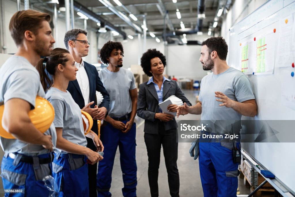 Young worker giving presentation in front of whiteboard in a factory. Young factory worker holding presentation about production development to company managers and his coworkers. Occupation Stock Photo