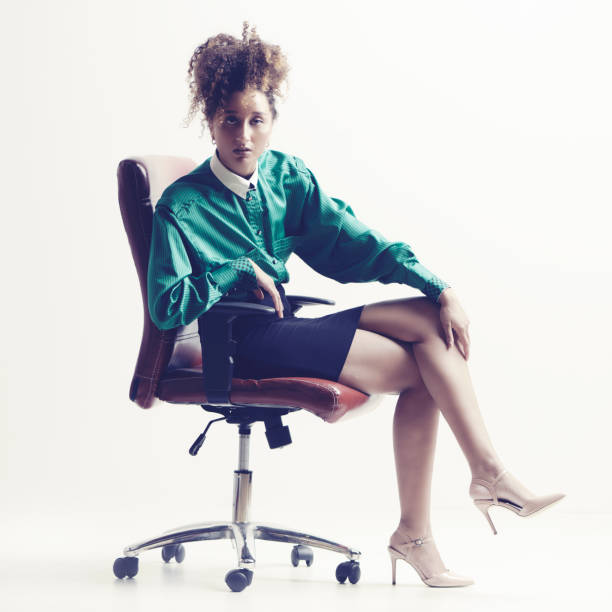 Office woman on chair. Side view of woman on chair with legs crossed at knee.  Ethnic mixed race woman on gray background in studio, desaturated  image with copy space