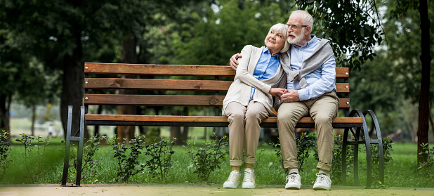 happy senior couple hugging while sitting on wooden bench in park