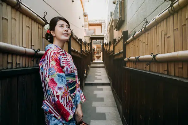japanese teenage girl wearing floral traditional dress standing in little path between building with bamboo wall. young elegant woman smile look up to blue sky in colorful kimono cloth in kyoto japan