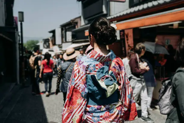 back view of japanese young girl in flower kimono dress walking in kiyomizu zaka street. woman join festival in old teeming town in kyoto japan. tourist sightseeing experience japanese culture.