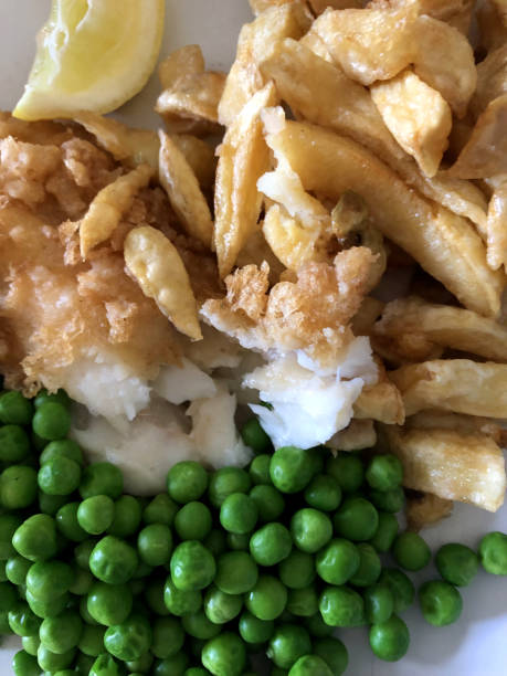 Stock photo of takeaway fish and chips with slice of lemon, garden peas / frozen peas, salt and vinegar seasoning, unhealthy food takeout, greasy battered cod and deep fried chips as English fish supper ready to eat for lunch at the seaside imagen de pescado para llevar y papas fritas con rebanada de limón, guisantes de jardín/guisantes congelados, condimento de sal y vinagre, comida saludable para llevar, bacalao maltrecha grasoso y papas fritas fritas como cena de pescado inglés listo pa