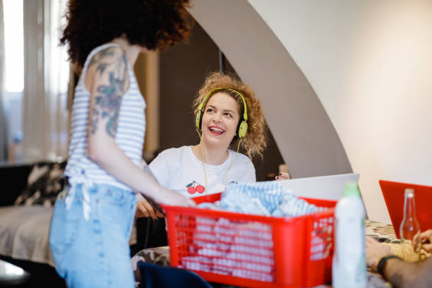 One woman is working at home on laptop while flatmate is doing chores Smiling young woman with headphones working at home on laptop while flatmate is doing chores flatmate stock pictures, royalty-free photos & images