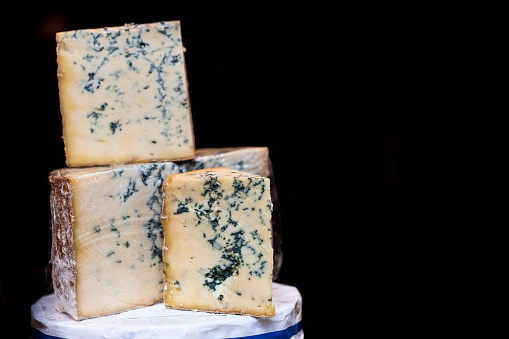 Horizontal color image depicting fresh British stilton cheese (a blue cheese) on display and for sale at Borough Market, London, UK. Focus is on three wedges of cheese in the foreground, while the background is entirely black. Room for copy space.