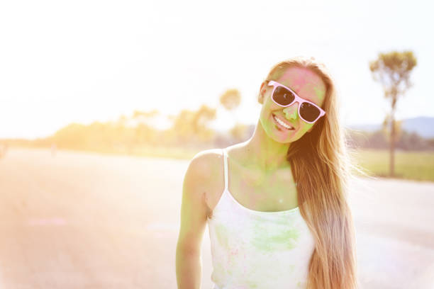 Woman loving the color festival, laughing and looking at camera at public park. Young candid girl with face paint wearing pink sunglasses and enjoying the event.