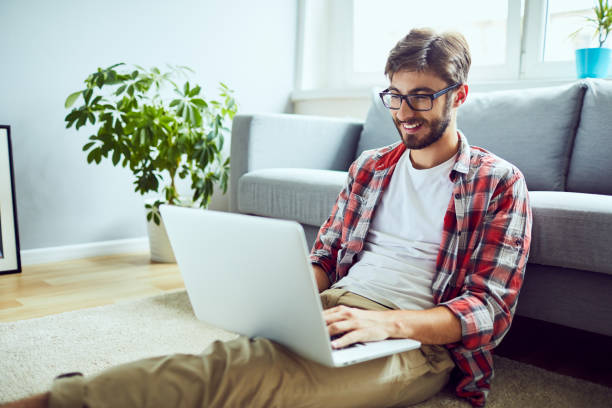 jeune homme détendu travaillant sur l’ordinateur portatif tandis qu’assis sur le plancher à la maison - smart casual cheerful content image photos et images de collection