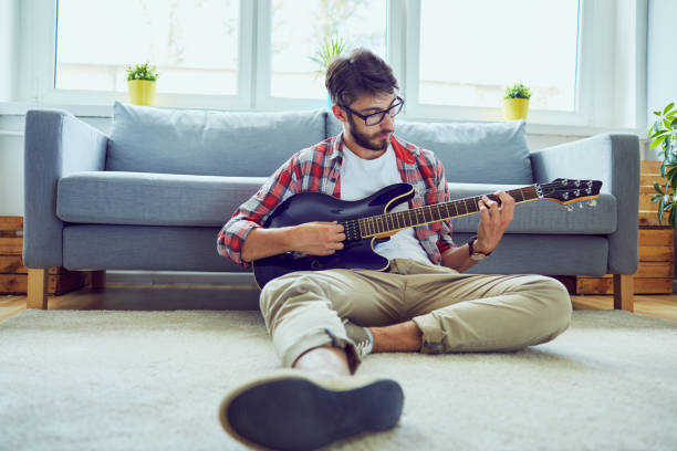 front view of a young handsome man playing the guitar while sitting on floor in his living room - guitarist one person caucasian adult imagens e fotografias de stock