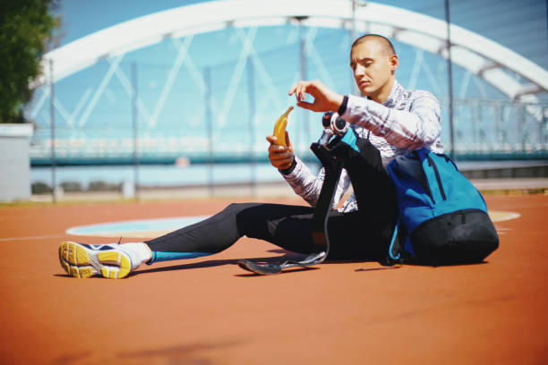 Young male athlete with artificial leg  taking a break at stadium track, sitting and eating banana.