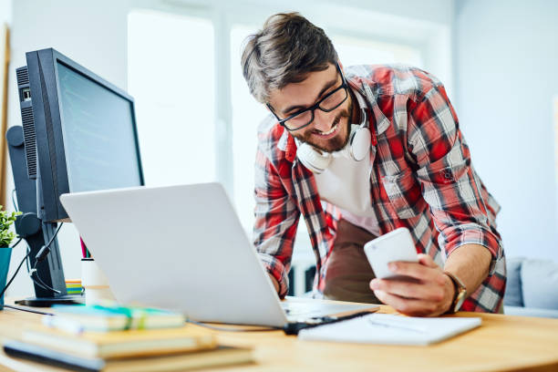 istock Smiling programmer standing at desk working on laptop and checking phone 1151965077