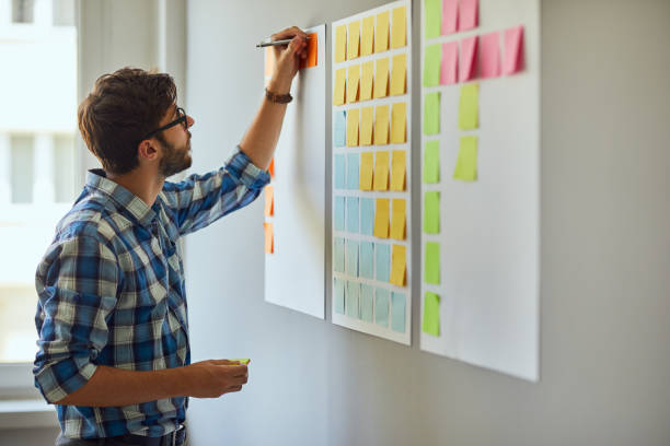 Young creative man writing down ideas on wall full of sticky notes stock photo