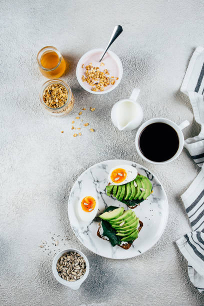 istock Healthy Breakfast concept. Sandwich with avocado, spinach, and boiled eggs, yogurt with granola, cup of coffee over concrete background, top view. Health, weight loss, detox, vegetarian, diet food concept 1151980134