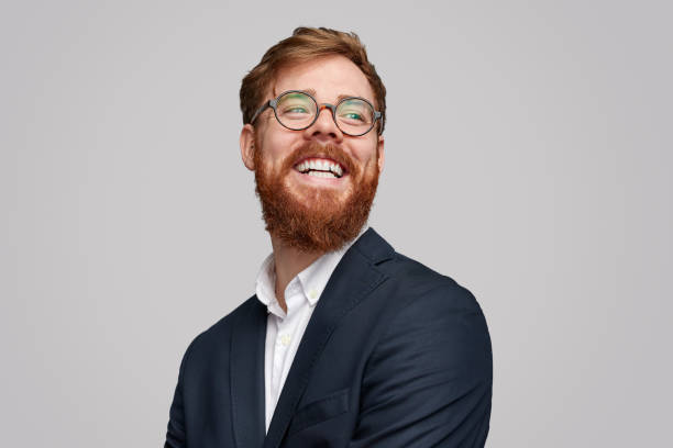 Cheerful red bearded businessman looking away Handsome Irish businessman with ginger beard wearing formal suit and glasses and cheerfully smiling isolated on gray background and looking away looking away stock pictures, royalty-free photos & images