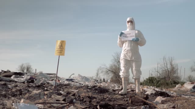 ecological disaster, woman in protective suit and mask holding poster with stop pollution slogan at landfill near sign biological hazard
