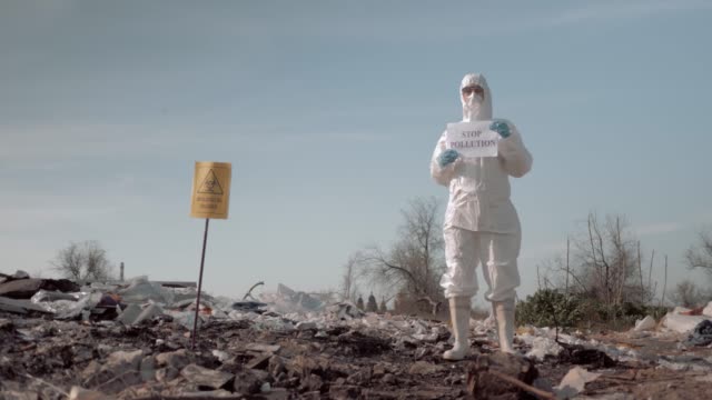 woman in protective suit and mask holding poster with stop pollution slogan at garbage dump near sign biological hazard
