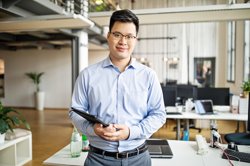 Portrait of mid adult asian businessman standing in office with digital tablet in hand. Male professional looking at camera while standing by a desk.