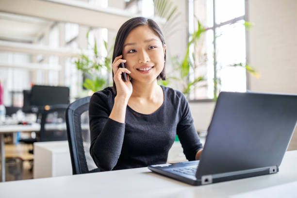 Female professional working in modern office Young asian businesswoman sitting at her desk with laptop talking on mobile phone. Female professional working in modern office. phone speak stock pictures, royalty-free photos & images