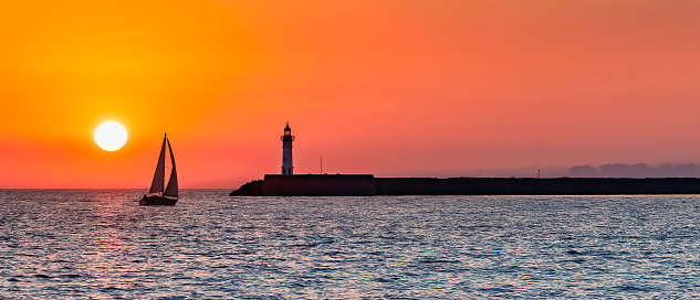 Beautiful panoramic view of an ocean sunset, with silhouettes of sail boat and lighthouse against an orange sky.