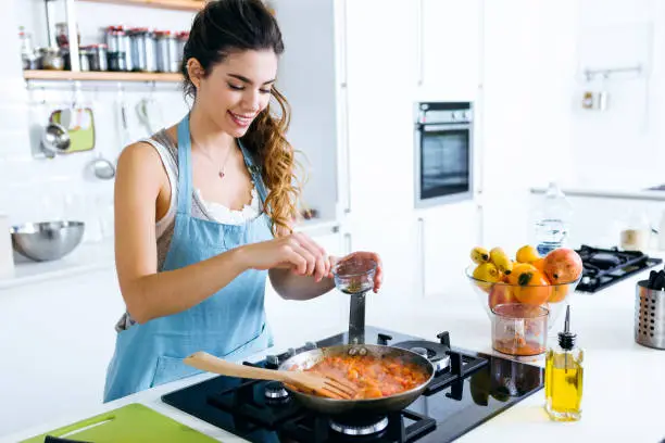 Photo of Young woman adding pepper and mixing food in frying pan.