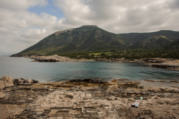 Three people in sportswear on rocky seashore looking at great green mountains in Cyprus