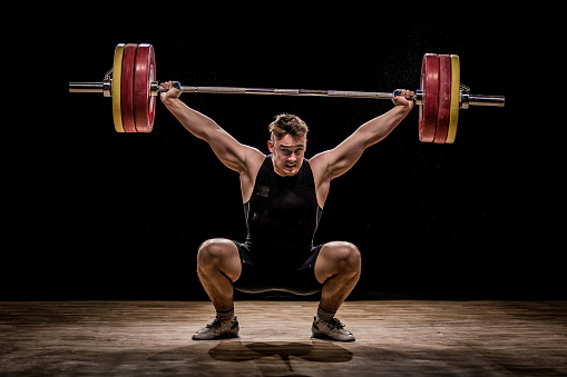 Crouching body builder holding barbells performing deadlift in front of a black background.