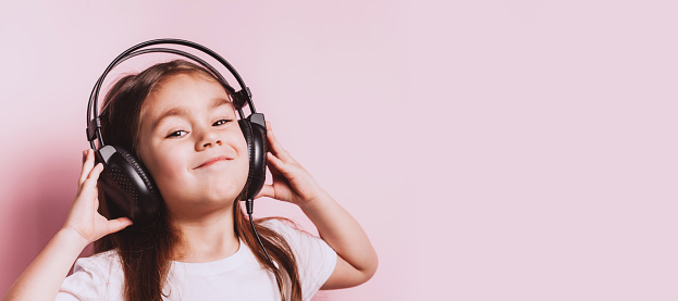 Cute little girl listening music wearing earphones on pink background. Funny emotions. Copyspace for text.