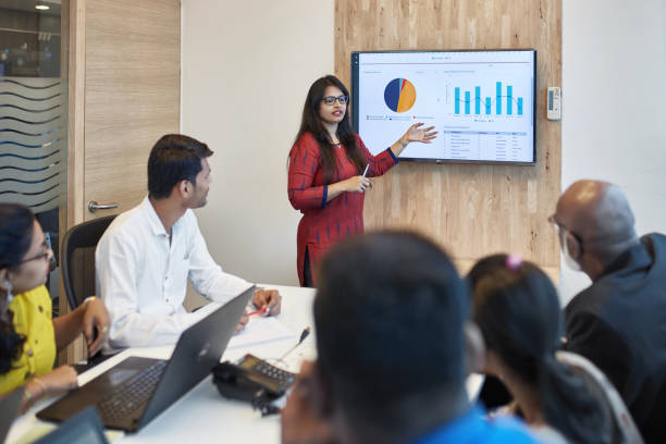 Businesswoman explaining graph to colleagues Confident young businesswoman explaining graph to professionals in board room. Colleagues are planning strategy at creative workplace. They are working at office. Plan stock pictures, royalty-free photos & images
