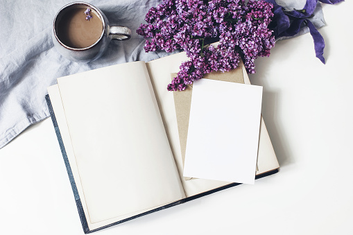 Spring feminine scene, floral composition. Bunch of purple and white lilac flowers, old book, cup of coffee and linen tablecloth on white background. Blank greeting card mockup, flat lay, top view.