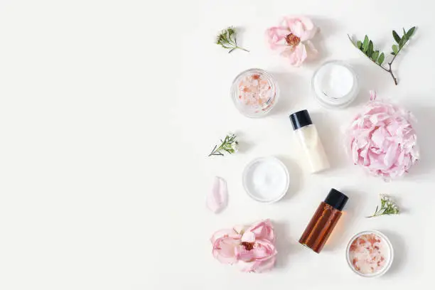 Styled beauty composition. Skin cream jar, tonicum bottle, roses, peony flower and Himalayan salt on white table background. Organic cosmetics, spa concept, empty space, flat lay, top view.