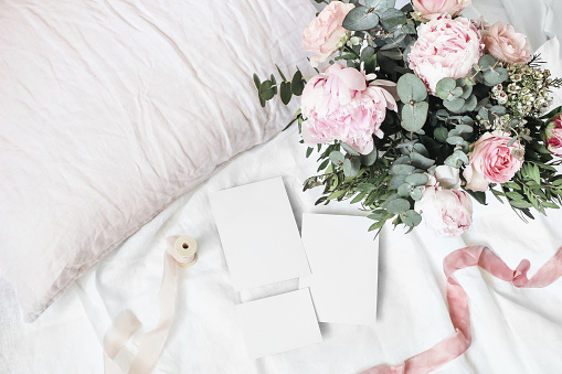 Cozy bedroom still life scene. Wedding, birthday bouquet of pink roses, peony flowers and eucalyptus branches. Silk ribbons on linen bedding. Blank greeting cards mockups, top view.