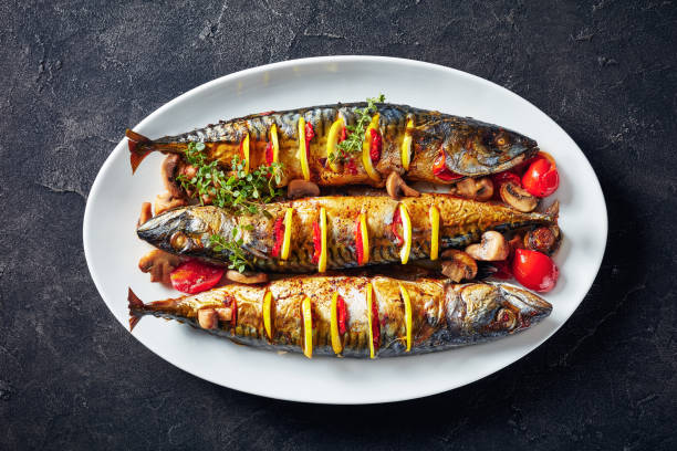 three whole grilled mackerel with lemon, tomatoes, mushrooms, spices and herbs. close-up of three whole broiled mackerels with lemon, tomatoes, mushrooms, spices and herbs on a white oval dish, horizontal view from above, flatlay, mackerel stock pictures, royalty-free photos & images