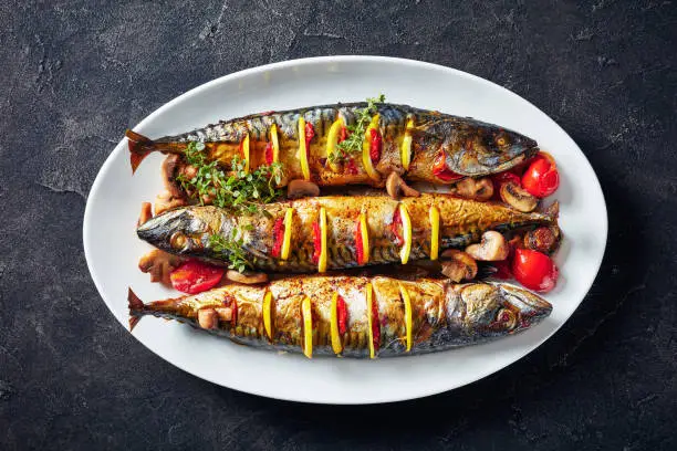 close-up of three whole broiled mackerels with lemon, tomatoes, mushrooms, spices and herbs on a white oval dish, horizontal view from above, flatlay,