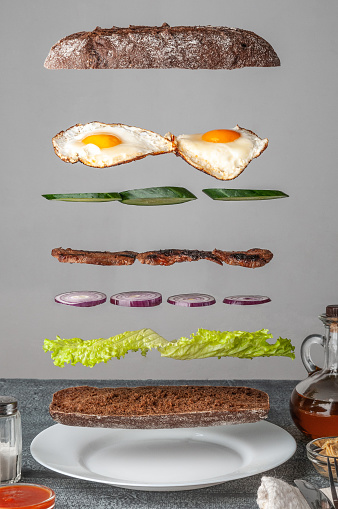 Sandwich ingredients are levitated in the air with crispy baguette, lettuce, cucumber, red onion, grill meat and scrambled eggs. In the foreground are cutlery, spices, sauce and butter. The background is gray.