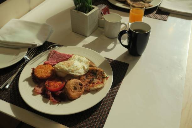 Stock photo of mixed full English Indian breakfast, starting the day with an egg and typical greasy fry up, indoors breakfast dining table eating in morning, New Delhi hotel, India. imagen de desayuno mixto inglés indio completo con huevo frito, jamón, tomates a la plancha, tocino crujiente, salchichas, vadas, grasoso kochuri, patatas hash marrón foto/aloo tikki, mesa de restaurante con café y jugo de naranja, desayuno en el inter