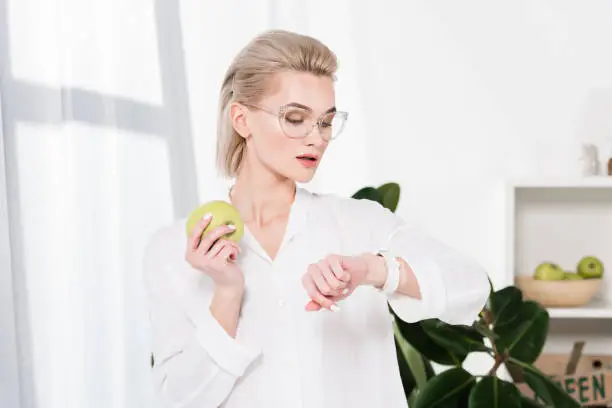 Photo of beautiful businesswoman in glasses looking at watch while holding green apple in office