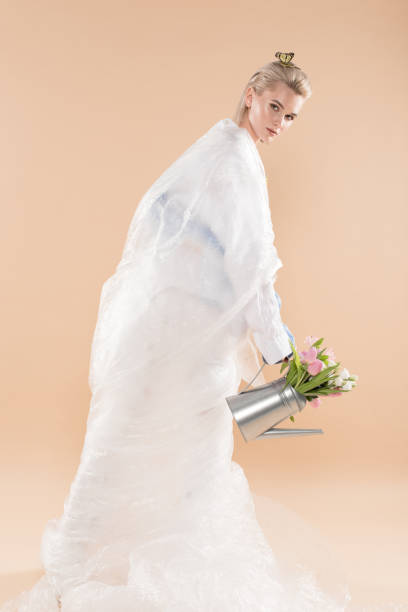 attractive toung woman wrapped in polyethylene holding watering can with flowers isolated on beige, environmental saving concept - toung imagens e fotografias de stock