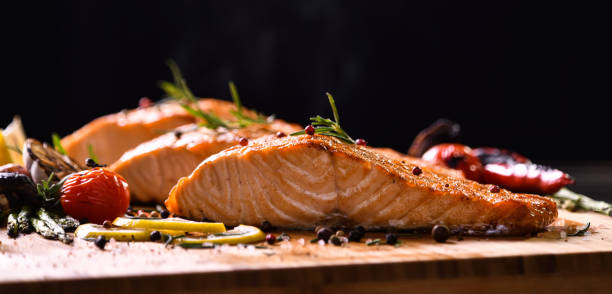 pesce salmone alla griglia e varie verdure su tavolo di legno su sfondo nero - prepared fish foto e immagini stock