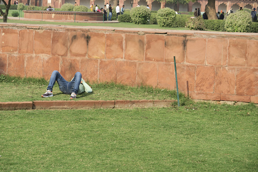 Stock photo showing a sleepy, tired Indian Hindu man who is sleeping in afternoon sunshine in lawn, sunbathing near Agra Fort, Uttar Pradesh, India, afternoon siesta hot weather, napping, snoozing, curled up sleeping on grass.