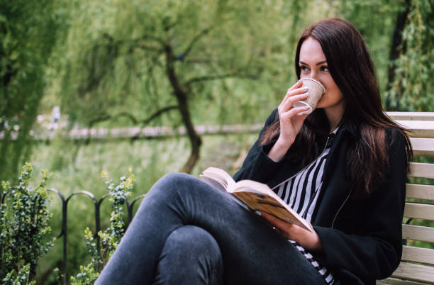 lächelnde brünette mädchen sitzen auf einer bank in einem park, wenn kaffee aus einer papierkasse trinken und in der ferne. weiches cover-buch in ihren händen. - shirt paper book business stock-fotos und bilder