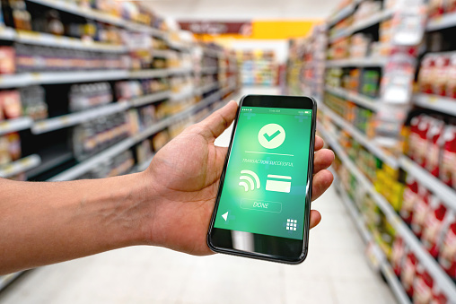 Unrecognizable person holding smartphone while making a transfer to pay for groceries at supermarket - Focus on foreground **DESIGN ON SCREEN WAS MADE FROM SCRATCH BY US**