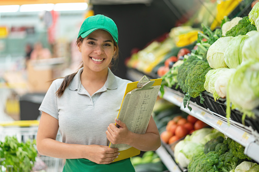 Latin american confident sales woman holding a tablet facing camera smiling at the vegetable section in supermarket