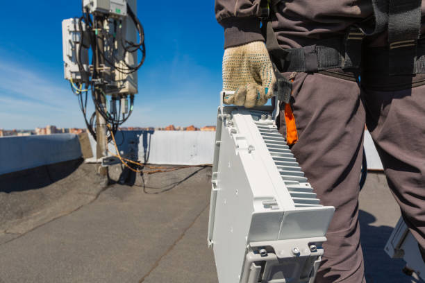 istock Telecommunication engineer  in helmet and uniform holds telecomunication equipment in his hand and  antennas of GSM  DCS UMTS LTE bands, outdoor radio units, optic fibers, power cables are installed on the roof. Working process of upgrading telecommunicat 1152074537