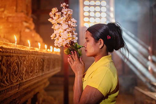 Burmese woman standing inside buddhist temple,praying to Buddha, holding a bouquet of flowers in her hands. Moody ambient natural light and light beams shining through the window of the temple pagoda.. Bagan, Mandalay, Myanmar, Asia.