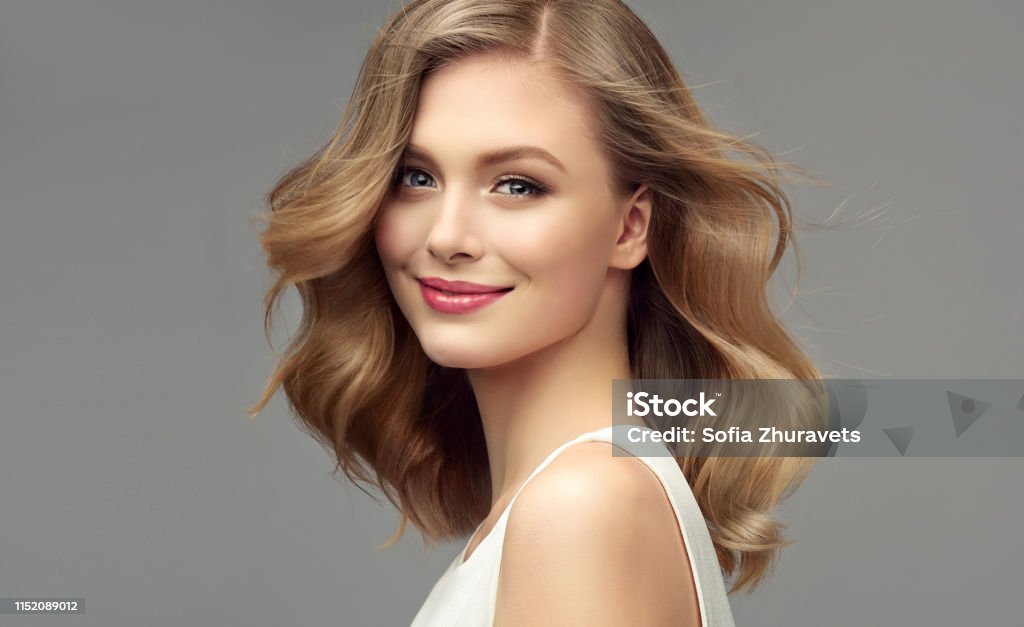 Portrait of young woman with dark blonde hair. Cosmetology, hairdressing and makeup. Model with dark blonde hair. Frizzy, elegant hairstyle is surrounding lovely face of tenderly smiling young woman. Natural gloss and softness of healthy hair. Hair care and hairdressing art. One Woman Only Stock Photo