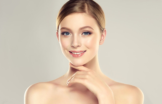 Gorgeous, young woman dressed in a soft, elegant makeup is touching the face by graceful fingers. Slender neck, bare silky shoulders and elegant gesture of hand. Facial treatment, cosmetology, beauty technologies and spa.